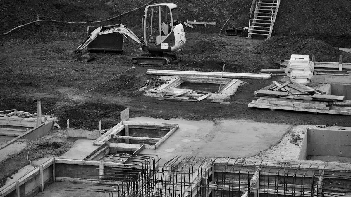 Monochrome image of an excavator at a construction site with foundations laid and rebar in place.